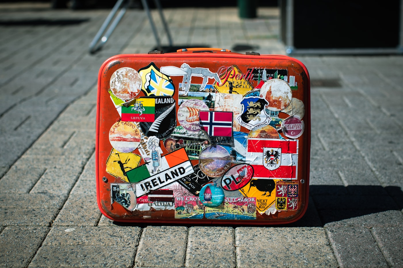 各国の観光ステッカーがびっしり貼られた赤いハードケースのスーツケースが石畳の上に置かれている様子